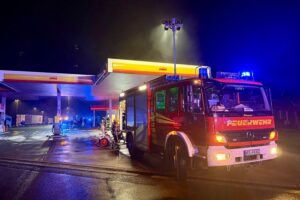 Brennender Pkw an Tankstelle, Feuerwehr im Einsatz, Nachtaufnahme, hohe Rauchentwicklung, dramatische Szene.