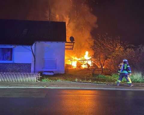 Ein abgebrannter Holzschuppen hinter einem Wohnhaus, Feuerwehr im Einsatz, Rauch und Zerstörung.