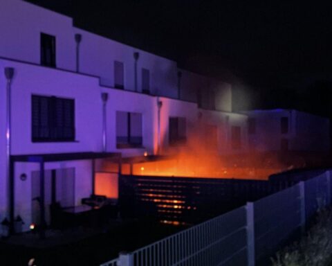 Feuerwehr löscht Brand in Reihenhaus; Flammen, beschädigte Fassade und zerbrochene Fenster sichtbar.
