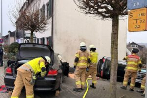 Unfallstelle mit schwarzem BMW, beschädigter Hauswand und Einsatzkräften der Feuerwehr.