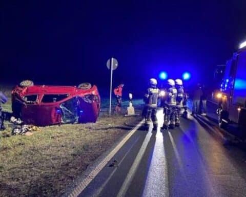 Unfallstelle mit beschädigtem Auto, Polizei und Rettungsdienst, Warnlichter, verletzte Personen am Boden.