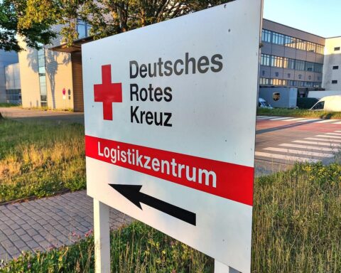 Ein DRK-Logistikzentrum mit Spendenboxen und hilfsbedürftigen Menschen im Hintergrund.