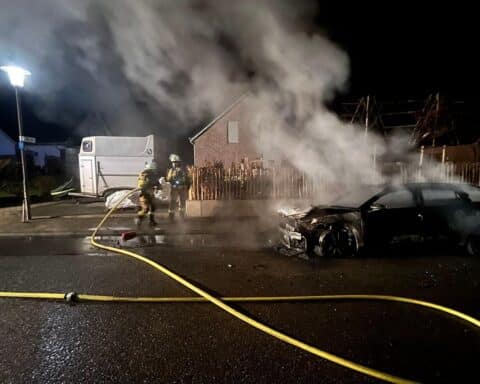 Ein brennender PKW auf einer Straße, Einsatz der Feuerwehr im Hintergrund.