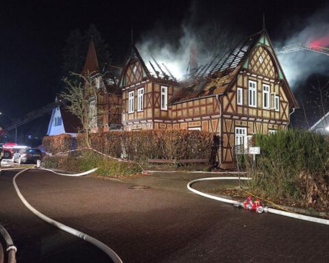 Abgebranntes Fachwerkhaus mit Feuerwehr im Einsatz, dichte Rauchwolken und zerstörtes Dach sichtbar.