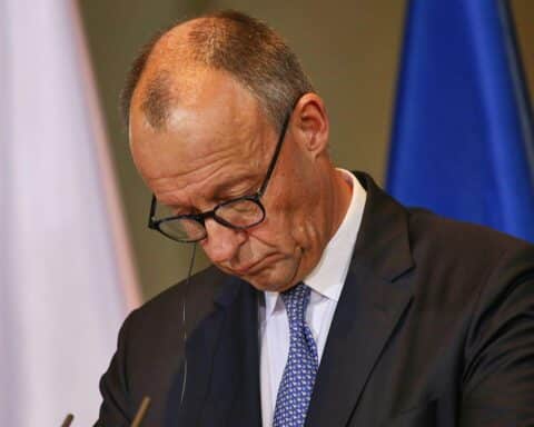 Friedrich Merz bei Pressekonferenz, im Hintergrund Wahlumfragen und politische Grafiken zu sehen.