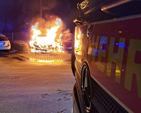 Brennendes Auto auf Parkplatz, Feuerwehr im Einsatz, dunkler Nachthimmel, dramatische Szene.