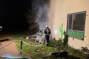 Einsatzkräfte der Feuerwehr löschen Brand, während Silvesterfeuerwerke im Hintergrund explodieren.