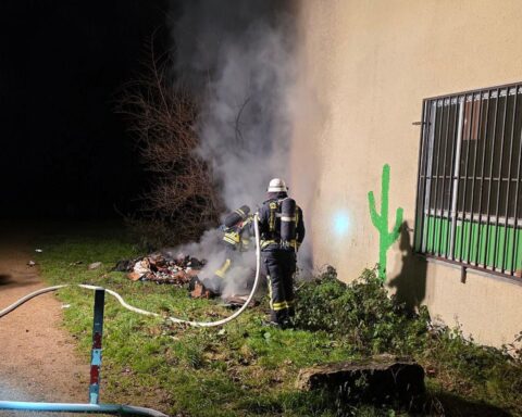 Einsatzkräfte der Feuerwehr löschen Brand, während Silvesterfeuerwerke im Hintergrund explodieren.