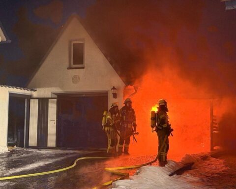 Einsatzkräfte löschen brennendes Auto unter Carport; Feuerwehr schützt angrenzende Gebäude vor Feuerausbreitung.