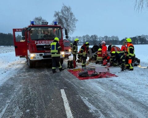 Feuerwehr und Rettungsdienst im Einsatz an Unfallstelle; schwer beschädigtes Fahrzeug unter Baum.