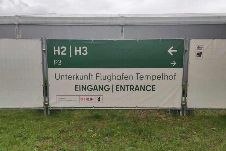 Flüchtlingsunterkunft mit Menschen, die auf Abschiebungen warten, europäische Flaggen im Hintergrund.
