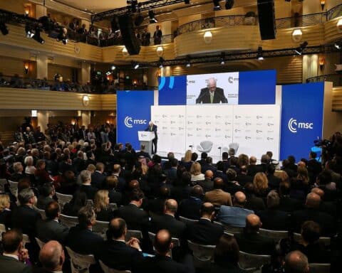 Politiker diskutieren Sicherheitsfragen; Demonstrationen im Hintergrund zeigen Bürgerengagement und aktuelle globale Themen.