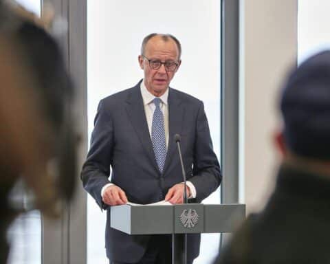 Friedrich Merz spricht mit angespanntem Gesicht, umgeben von kritischen Politikern vor einem diplomatischen Hintergrund.