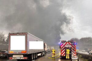 Brennender Lastwagen auf Autobahn, Feuerwehr im Einsatz, Rauch und Verkehrsbehinderungen sichtbar.