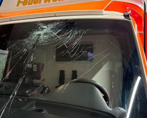 Zerstörte Windschutzscheibe eines Rettungswagens, umgeben von Polizei und ermittelnden Beamten.