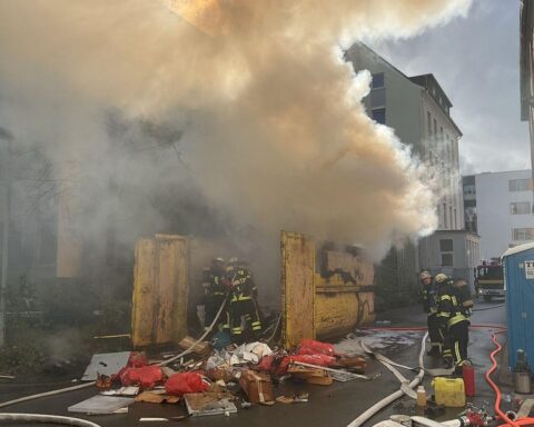 Feuerwehr löscht brennenden Müllcontainer nahe Kliniken, starke Rauchentwicklung und Gefahr für Gebäude.