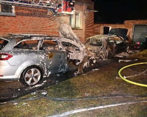 Zwei vollständig ausgebrannte Autos vor einem Mehrfamilienhaus, Feuerwehr im Einsatz.