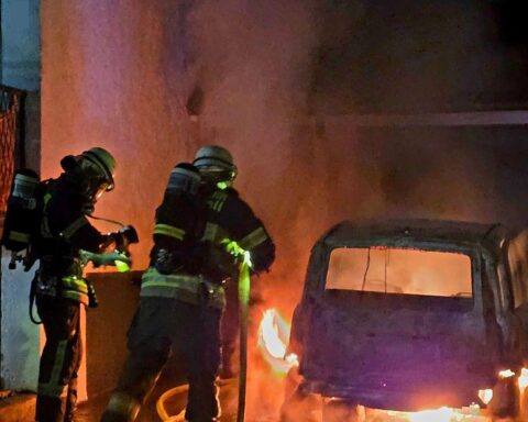 Ein ausgebranntes Auto vor einem Wohngebäude, Feuerwehrleute löschen den Brand.