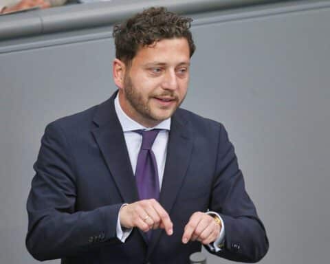 Felix Banaszak kritisiert Kanzler Merz' Krisenmanagement in Berlin, symbolisiert durch eine Pressekonferenz.