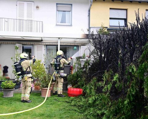Brennende Hecke zwischen Reihenhäusern, Feuerwehr im Einsatz, Rauch und Flammen sichtbar.