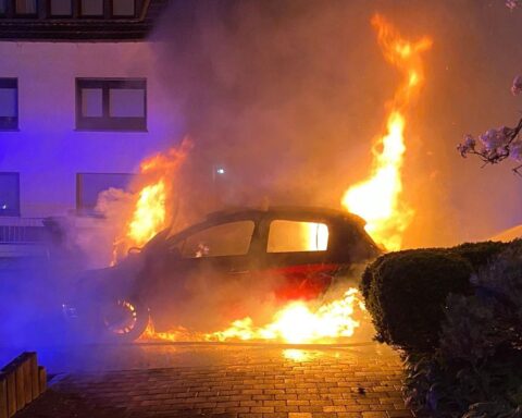 Ein brennendes Elektroauto am Straßenrand, Feuerwehr löscht, Rauch und Flammen sichtbar.