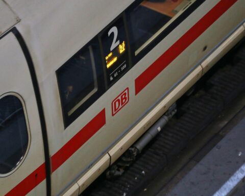 Eisenbahn-Zug im Hintergrund, Fahrgäste warten ungeduldig, Preisschilder deuten auf steigende Kosten hin.