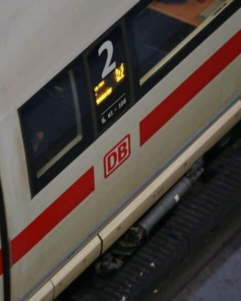 Eisenbahn-Zug im Hintergrund, Fahrgäste warten ungeduldig, Preisschilder deuten auf steigende Kosten hin.