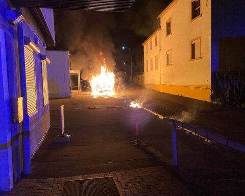 Ein brennendes Auto steht in Flammen, Rauch und Zerstörung im Hintergrund sichtbar.
