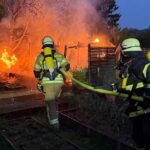 Feuerwehr löscht Brand in Gartenhütte; Rauchwolke und Einsatzkräfte im Einsatz sichtbar.