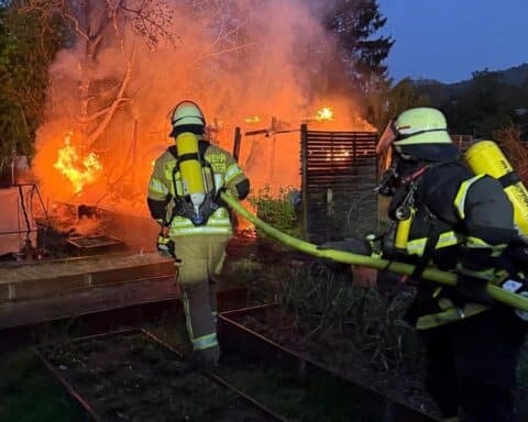 Feuerwehr löscht Brand in Gartenhütte; Rauchwolke und Einsatzkräfte im Einsatz sichtbar.