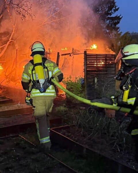 Feuerwehr löscht Brand in Gartenhütte; Rauchwolke und Einsatzkräfte im Einsatz sichtbar.