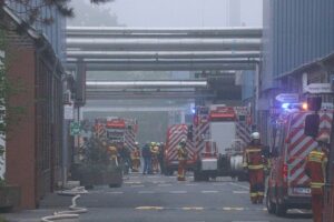 Feuerwehrleute löschen Brand in Industriebetrieb, Rauch und Flammen auf Dach und Rohrleitungen sichtbar.