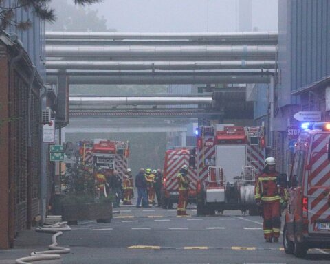 Feuerwehrleute löschen Brand in Industriebetrieb, Rauch und Flammen auf Dach und Rohrleitungen sichtbar.