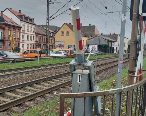 Bahnübergang mit beschädigter Schranke, prominent platzierten LKW und darauf hinweisenden Verkehrsschildern.