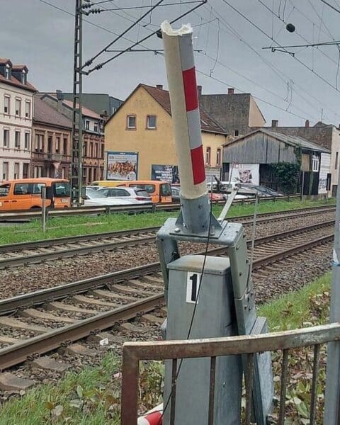 Bahnübergang mit beschädigter Schranke, prominent platzierten LKW und darauf hinweisenden Verkehrsschildern.