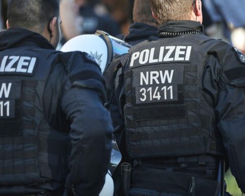 Ein Polizist untersucht eine blutige Tatstelle mit verletztem Mann und Ermittlern in der Nähe.