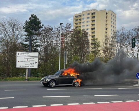 Brennender VW Käfer auf der Straße, Feuerwehr im Einsatz, starke Rauchentwicklung sichtbar.