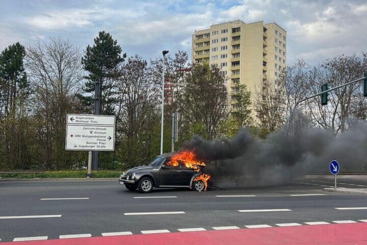 Brennender VW Käfer auf der Straße, Feuerwehr im Einsatz, starke Rauchentwicklung sichtbar.