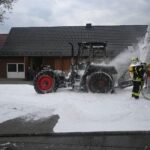 Traktorbrand auf Straße, Feuerwehr im Einsatz, Qualm und Flammen sichtbar, Schadensmeldung.