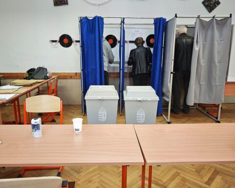 Wahllokal in Ungarn mit wartenden Wählern, farbigen Wahlurnen und Wahlplakaten im Hintergrund.