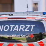 Unfallstelle mit zerstörtem Auto, Feuerwehrleuten und Notarzt im Einsatz zur Menschenrettung.
