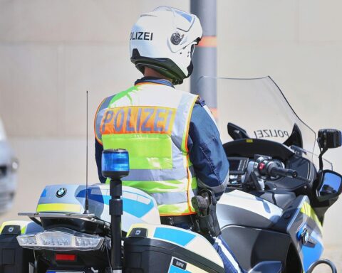 Motorrad-Polizist vor Unfallstelle mit BMW, verletztem Fahrer und nahegelegenen Kindern im Garten.