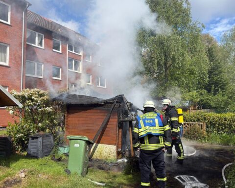 Holzschuppen brennt lichterloh vor Mehrparteienhaus, Feuerwehr im Einsatz, Flammen und Rauch sichtbar.