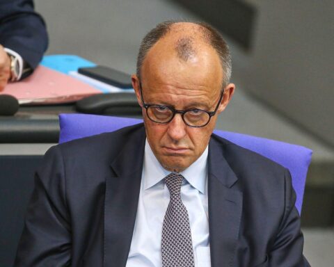 Friedrich Merz am Rednerpult, umgeben von politischen Symbolen und kritischen Zeitgenossen.