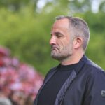 Manuel Baum lächelt, trägt FC Augsburg-Trainingsausstattung, umgeben von Co-Trainern in einem Stadion.
