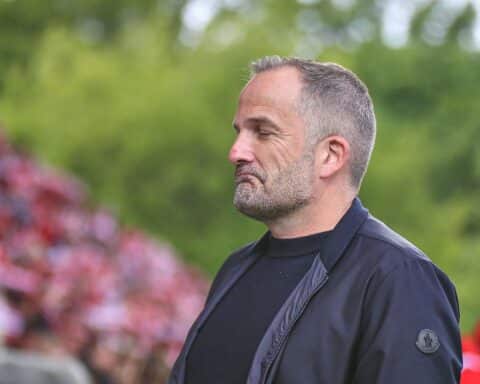 Manuel Baum lächelt, trägt FC Augsburg-Trainingsausstattung, umgeben von Co-Trainern in einem Stadion.