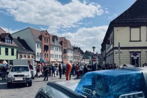 Friedliche Versammlung mit bunten Fahnen und Menschen, Polizei im Hintergrund überwacht das Geschehen.
