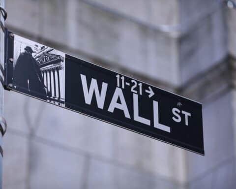 Straßenschild Wall Street mit Finanzsymbolik, glänzende Gebäude und Börsennachrichten im Hintergrund.