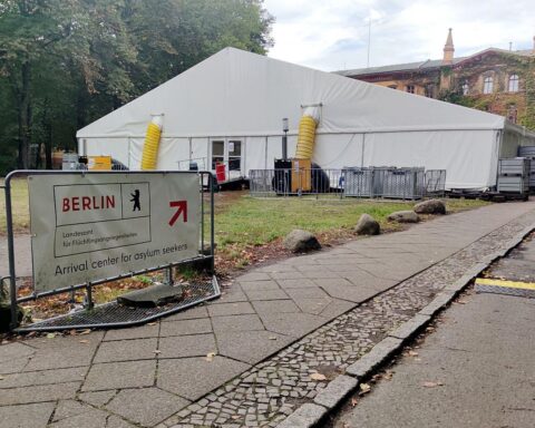 Ankunftszentrum für Flüchtlinge mit Symboliken zu Rückkehrprämien für Syrer und Integration.