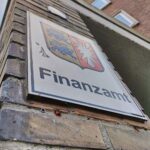 Ein Finanzamt-Gebäude mit einem Schild "Steuerreform" im Vordergrund und Menschen, die diskutieren.